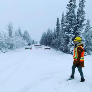 Photographie d’un chemin enneigé barré par des barrières. Une ingénieure en casque de chantier observe l’horizon.